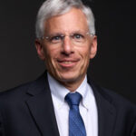 A professional headshot of Board member Bill Jeffrey in a black suit in front of a black background.