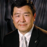 A professional headshot of Board member Bill Tong in a black suit in front of a grey background.