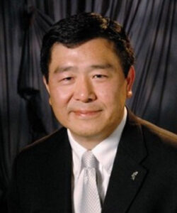 A professional headshot of Board member Bill Tong in a black suit in front of a grey background.