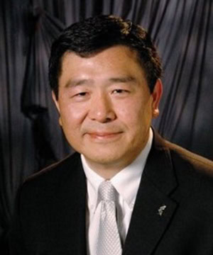 A professional headshot of Board member Bill Tong in a black suit in front of a grey background.
