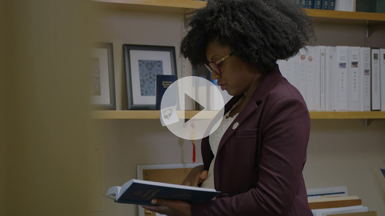 Debra Cooper in her office paging through a legal book with a play button overlaid to indicate it is a video link.