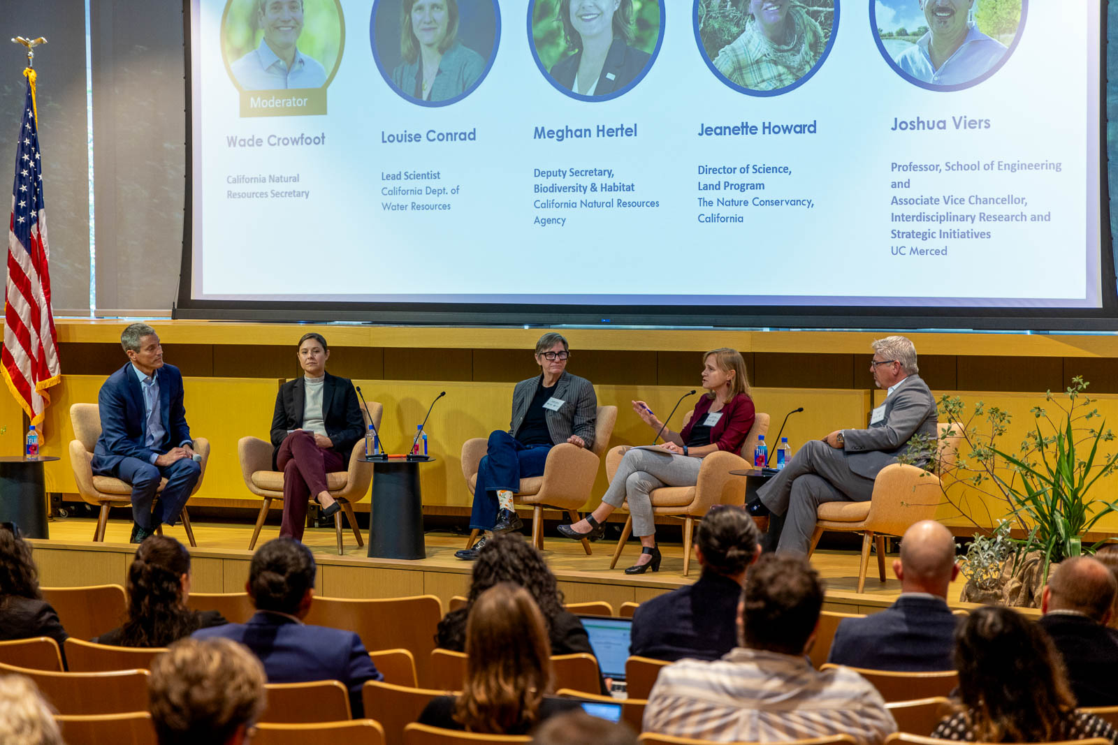 Panelists sit on a stage in front of a screen projecting their images, names, and titles.