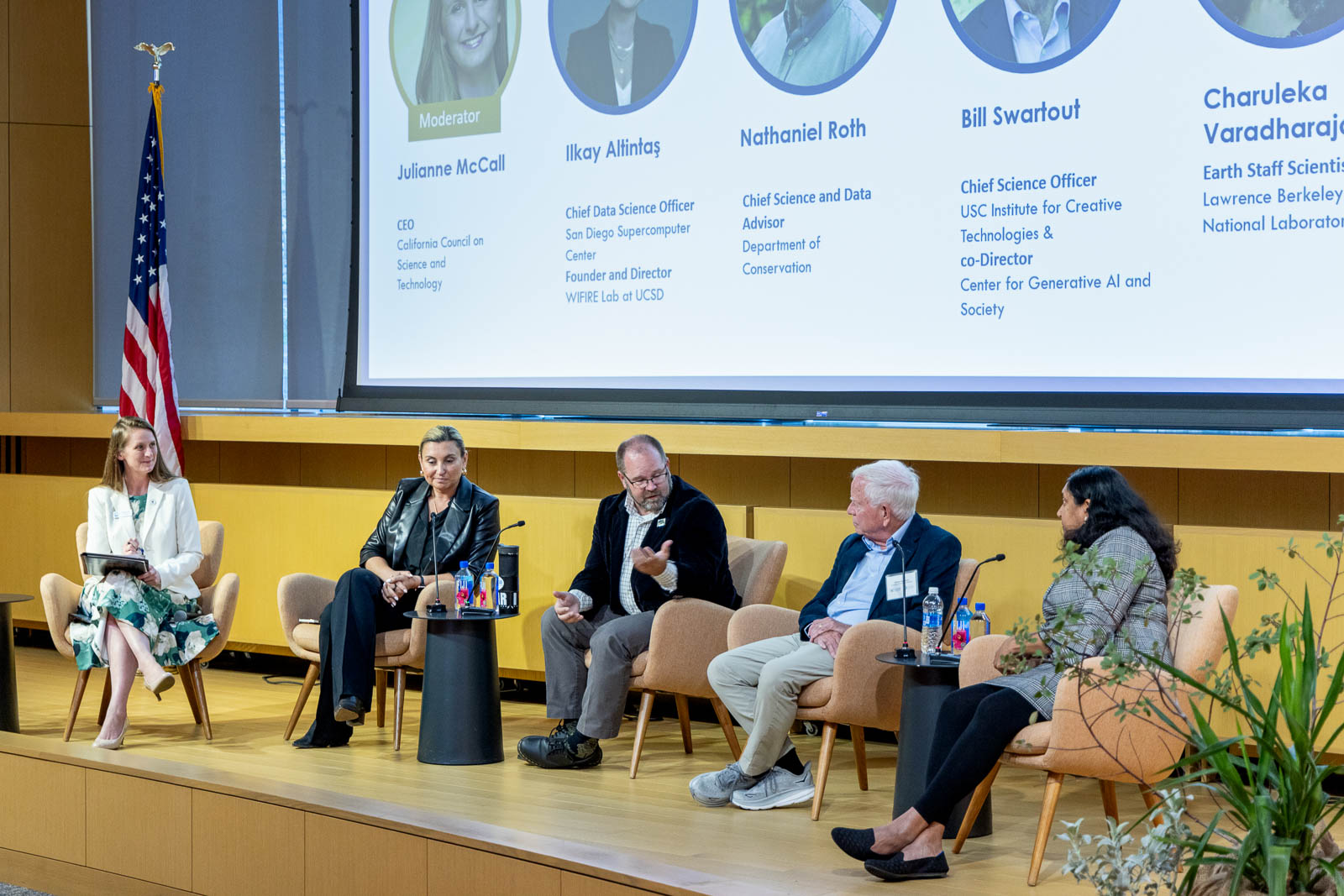 Panelists sit on a stage in front of a screen projecting their images, names, and titles.