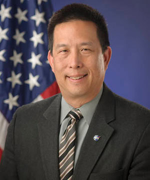 A professional headshot of the Board member with an American flag in the background.