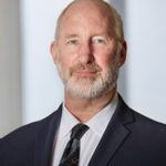 A professional headshot of Board member Glenn Fox in a suit in front of a light background.