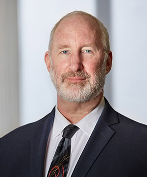 A professional headshot of Board member Glenn Fox in a suit in front of a light background.