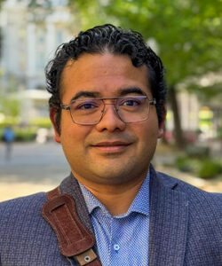 A headshot of Héctor Corzo in a suit outside with trees and the Capitol visible in the background