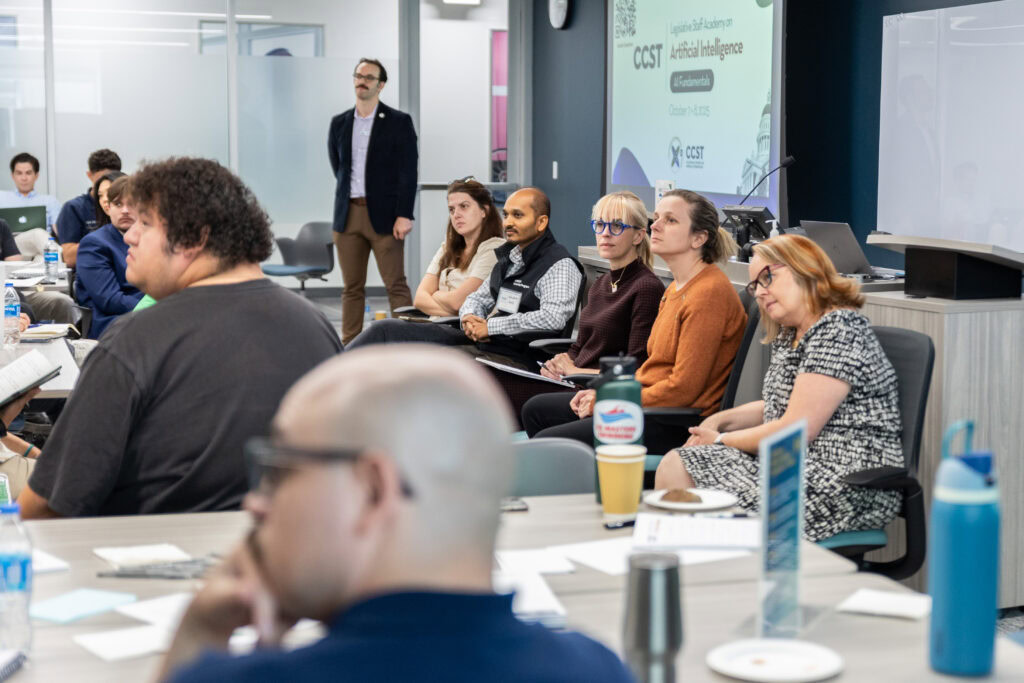 Five panelists lean forward listening to an audience question. Attendees visible in the foreground