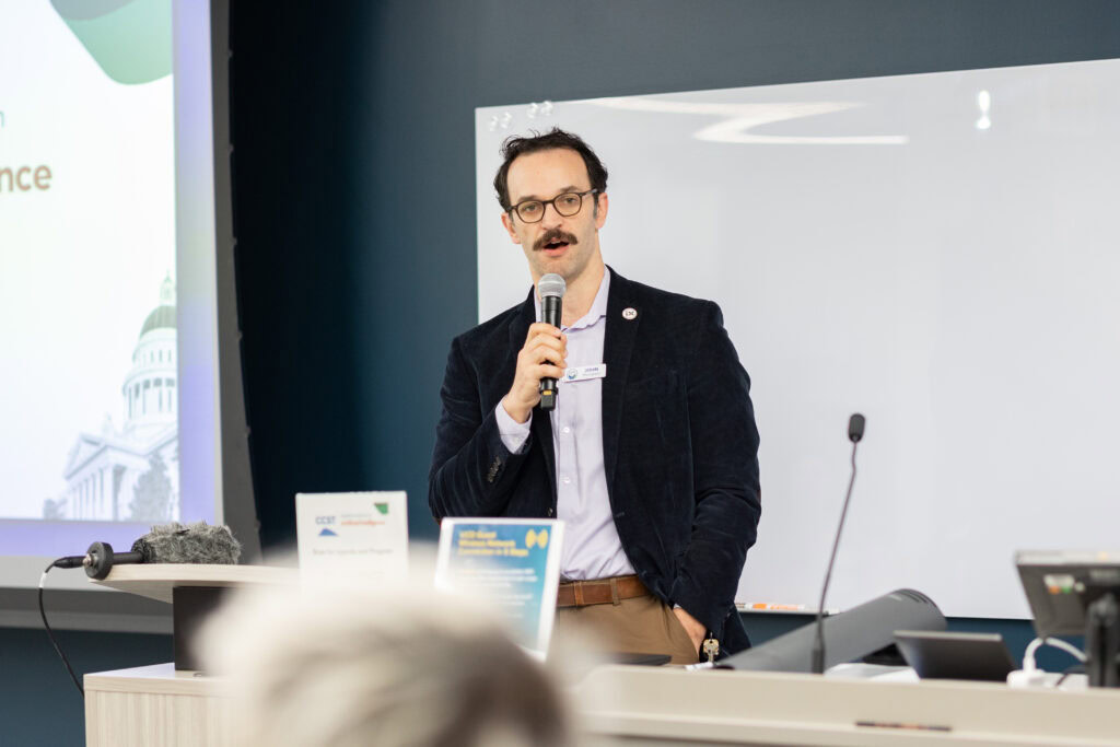 CCST Science Services Manager John Thompson, PhD, wearing a black suit jacket and light blue shirt, speaking into a microphone.