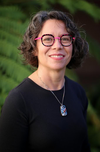 A professional headshot of Board member Rachel Segalman, smiling in a dark shirt with greenery in the background, blurred by the camera.