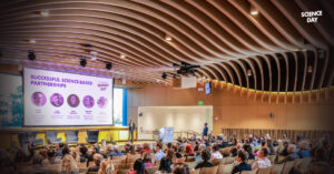 View of an auditorium with an audience and a projector screen featuring Science Day branding with Dr. Amber Mace on stage.