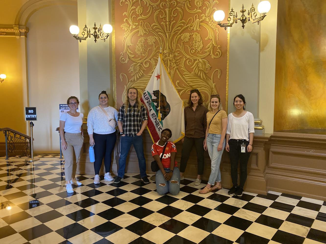Seven CCST fellows posing together for a photo inside the California State Capitol in front of the state flag on a black and white checkerboard floor.