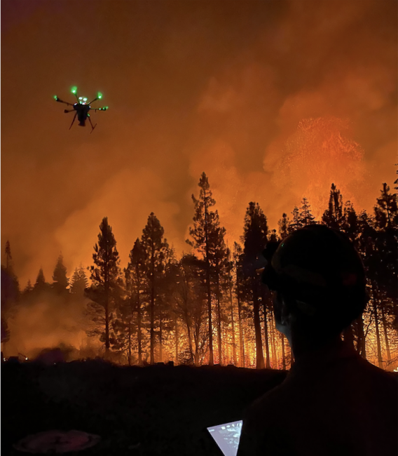 A drone flies above a wildfire landscape with the drone operator visible in the foreground.
